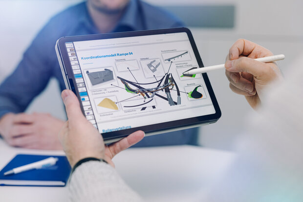 Eine Person hält mit einer Hand einen Tablet-Computer, auf dem Auszüge aus einem digitalen Modell erkennbar sind, und bearbeitet diese mit einem Touch Screen Stift. Im Hintergrund ist eine zweite Person zu erkennen.