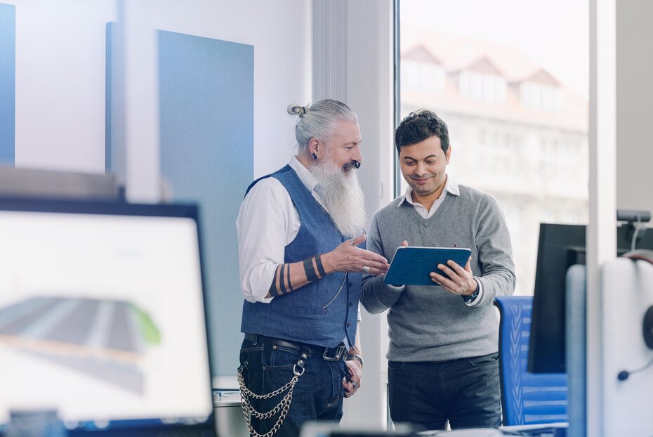 Ein älterer und ein jüngerer Mann blicken gemeinsam auf einen Tablet-PC. Sie sind umgeben von weiteren Bildschirmen, von denen einer den Ausschnitt eines BIM-Modells zeigt. 