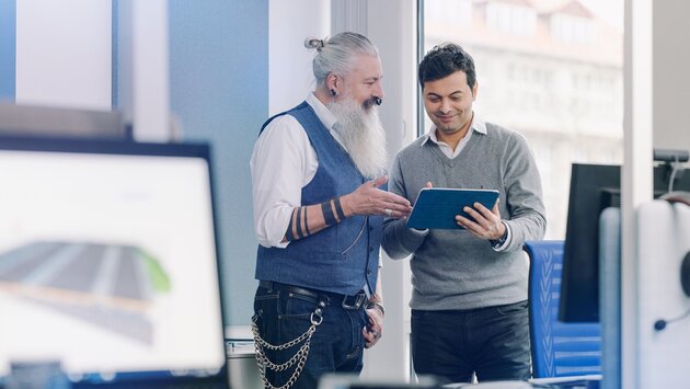 Zwei Männer stehen in einem Büroraum und unterhalten sich über Inhalte, die auf einem Tablet gezeigt werden, das einer der beiden Männer in der Hand hält.