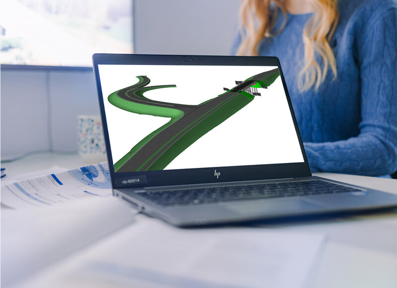 Ein Laptop steht auf einem Schreibtisch. Der Bildschirm zeigt eine BIM-generierte Darstellung einer Straße mit Unterführung. Im Hintergrund sind ein Büro, ein größerer Monitor an der Wand und eine Mitarbeiterin im blauen Pullover zu erkennen.