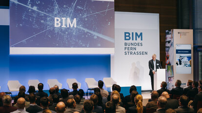 Starttrailer auf Leinwand, auf der groß und leuchtend die Buchstaben BIM zu sehen sind, Moderator Klaus Lintemeier am Pult, Publikum im Saal