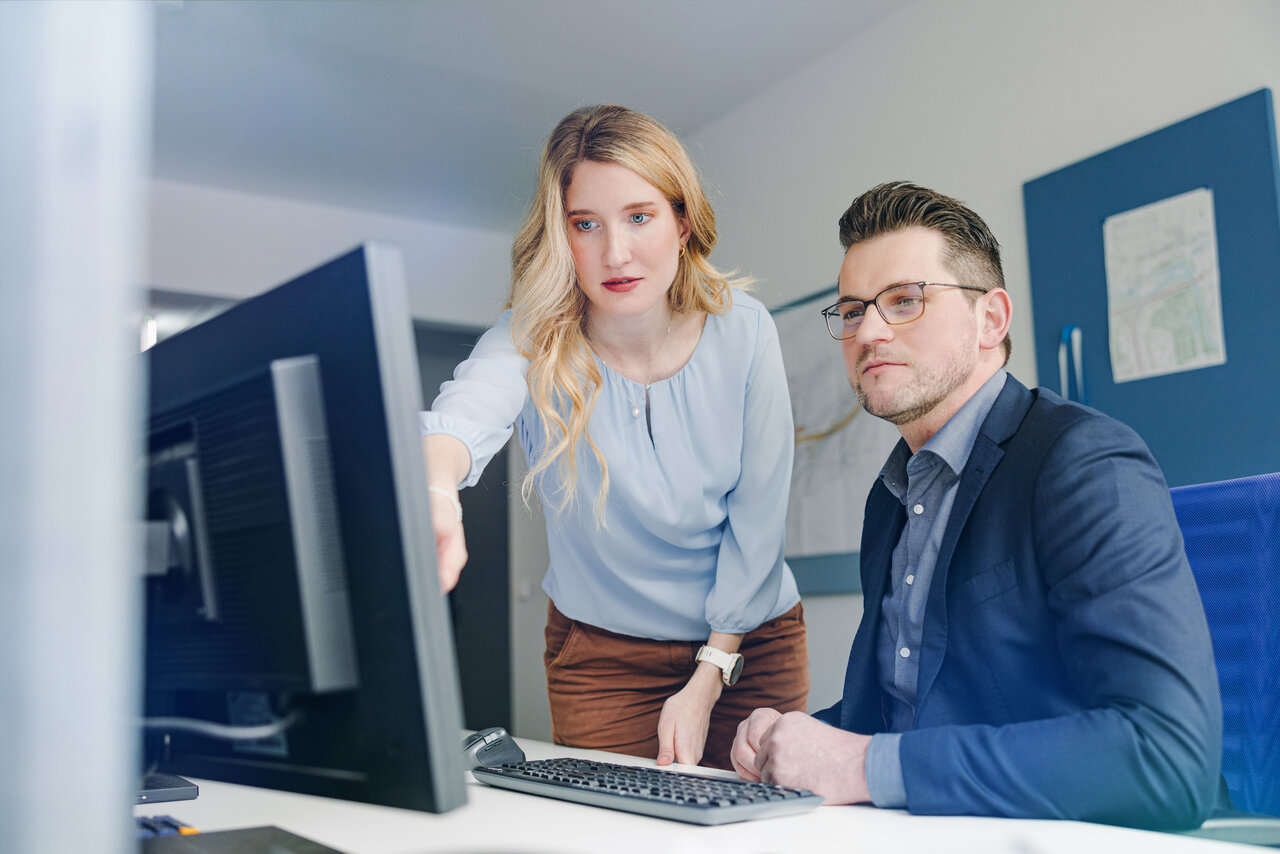 Zwei Personen im Büro schauen gemeinsam auf einen Computerbildschirm; die Frau zeigt auf den Monitor, der Mann sitzt daneben und hört aufmerksam zu. 